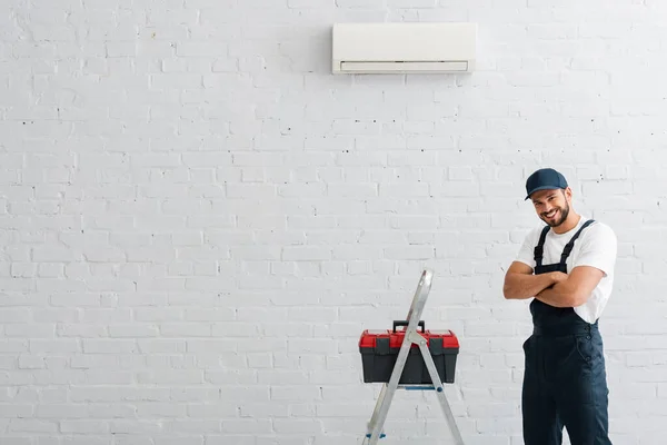 Trabajador sonriente con brazos cruzados mirando a la cámara cerca de la caja de herramientas en la escalera y aire acondicionado en la pared - foto de stock