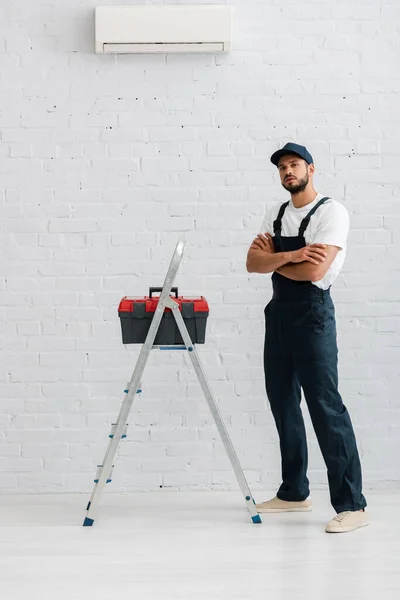 Handsome workman in overalls looking at camera near toolbox on ladder and air conditioner on white wall — Stock Photo