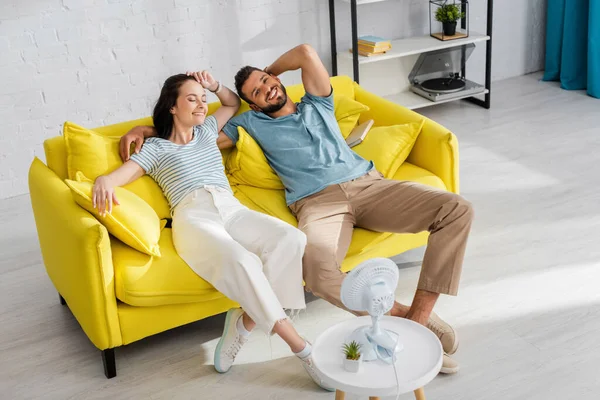 Selective focus of smiling couple sitting on sofa near electric fan on coffee table — Stock Photo