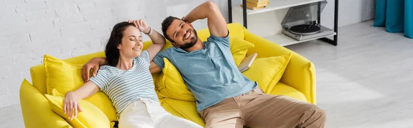 Récolte horizontale de bel homme souriant à la caméra près de copine attrayante sur le canapé à la maison — Photo de stock