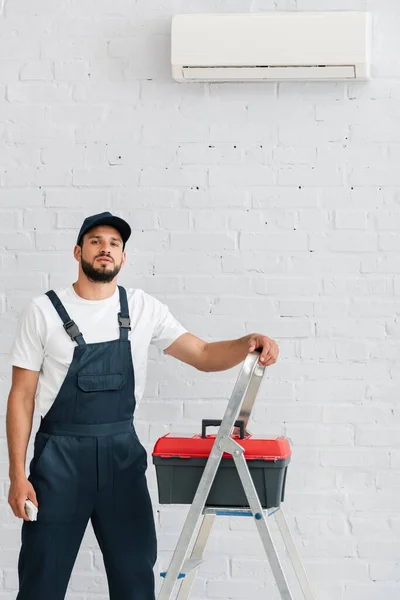Handsome workman looking at camera while holding remote controller of air conditioner near toolbox on ladder — Stock Photo