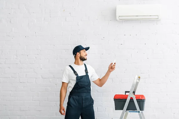 Lächelnder Arbeiter schaltet Klimaanlage mit Fernbedienung in der Nähe von Werkzeugkasten auf Leiter — Stockfoto