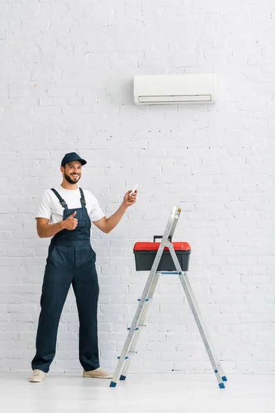 Trabajador sonriente mostrando el pulgar hacia arriba mientras sostiene el controlador remoto del aire acondicionado cerca de la caja de herramientas en la escalera - foto de stock