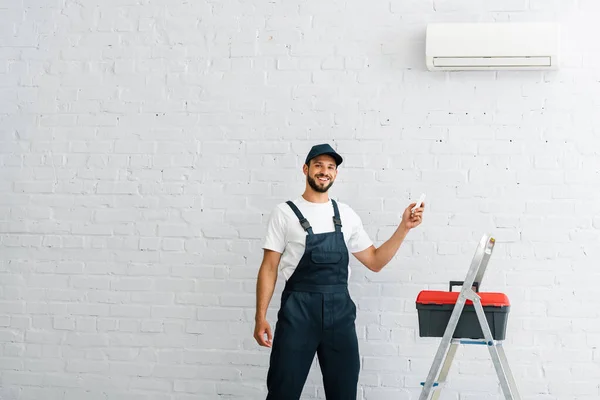 Schöner Arbeiter in Overalls lächelt, während er die Fernbedienung der Klimaanlage neben Werkzeugkiste und Leiter hält — Stockfoto