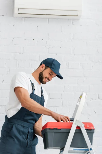 Lächelnder Arbeiter in Uniform hält Werkzeugkiste neben Leiter und Klimaanlage an weißer Wand — Stockfoto