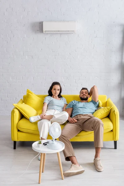 Foco seletivo do jovem casal olhando para a câmera perto do ventilador elétrico na mesa de café na sala de estar — Fotografia de Stock