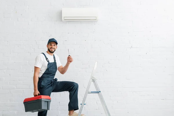 Trabajador sonriente con destornillador de sujeción uniforme cerca de escalera y aire acondicionado en pared - foto de stock