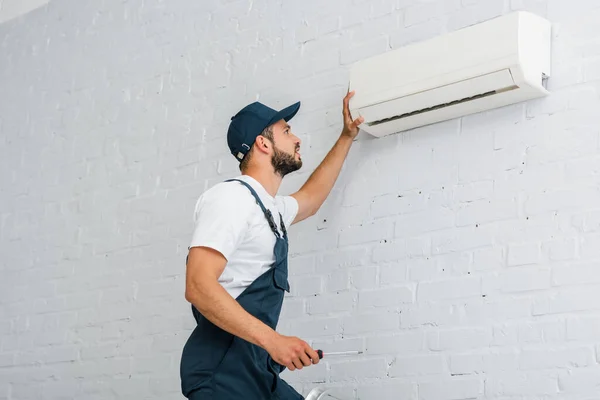 Side view of workman in workwear holding screwdriver near air conditioning — стоковое фото