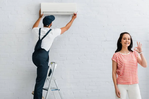 Donna sorridente mostrando gesto ok mentre riparatore fissaggio condizionatore d'aria — Foto stock