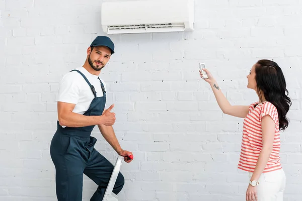 Obrero guapo mostrando el pulgar hacia arriba cerca de la mujer sonriente utilizando el controlador remoto de aire acondicionado - foto de stock