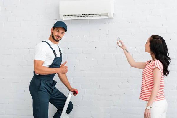 Beau travailleur montrant pouce vers le haut près de la femme souriante en utilisant la télécommande du climatiseur — Photo de stock