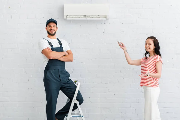 Homme à tout faire souriant regardant la caméra près de la femme pointant avec la main et tenant télécommande — Photo de stock