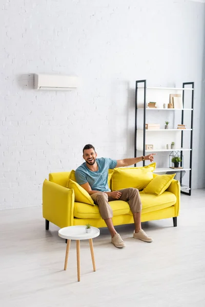 Hombre positivo señalando con el dedo y mirando la cámara en el sofá en casa - foto de stock