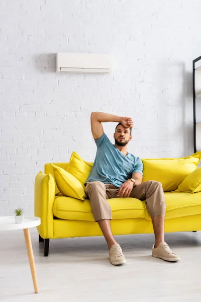 Selective focus of handsome man with hand near forehead feeling hot on couch at home — Stock Photo