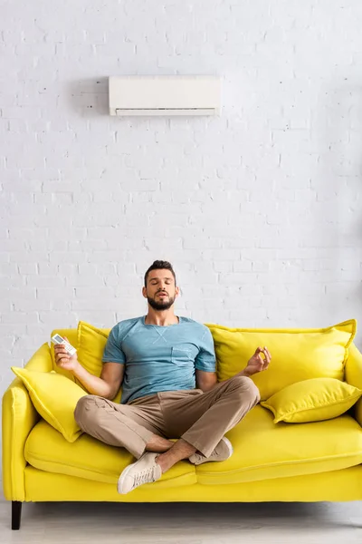 Hombre guapo meditando mientras sostiene el mando a distancia del aire acondicionado en el sofá - foto de stock