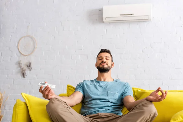 Hombre guapo sentado en postura de yoga mientras sostiene el controlador remoto de aire acondicionado en el sofá - foto de stock