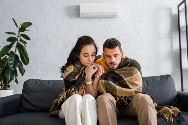 Casal jovem congelado embrulhando em xadrez no sofá em casa — Fotografia de Stock