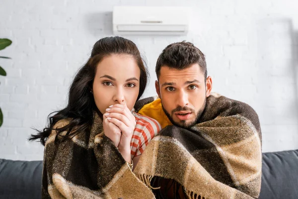 Jeune couple regardant la caméra tout en enveloppant dans plaid à la maison — Photo de stock