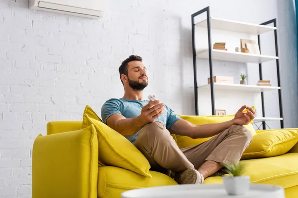 Selective focus of handsome man meditating while holding remote controller of air conditioner on sofa — Stock Photo