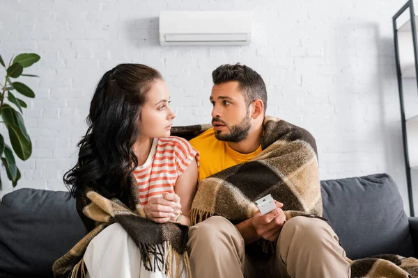 Jeune femme regardant petit ami enveloppé dans plaid et tenant télécommande de climatiseur sur le canapé — Photo de stock