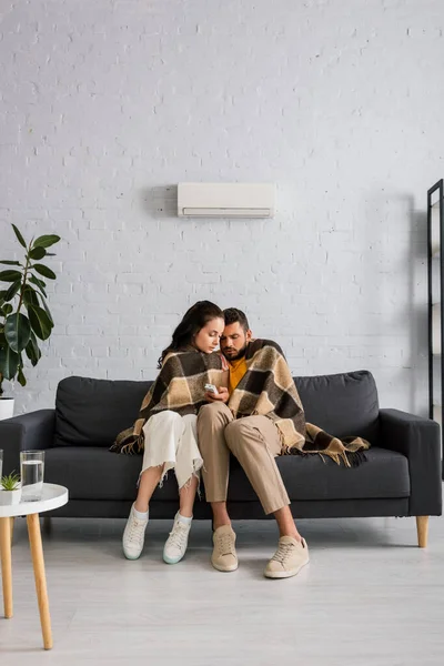 Couple enveloppant en plaid et tenant télécommande du climatiseur tout en se sentant froid sur le canapé — Photo de stock