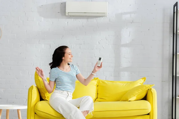 Sorrindo mulher segurando controlador remoto de ar condicionado no sofá em casa — Fotografia de Stock