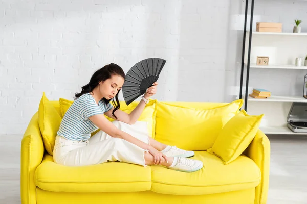 Vista lateral da menina cansada segurando fã enquanto se sente quente no sofá em casa — Fotografia de Stock
