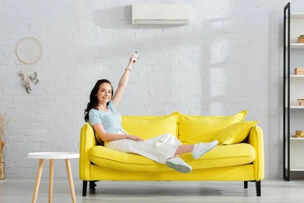 Hermosa chica sonriendo a la cámara mientras cambia el aire acondicionado con controlador remoto en casa - foto de stock