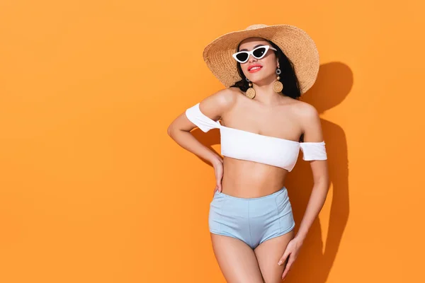 Chica feliz en traje de baño, gafas de sol y sombrero de paja de pie con la mano en la cadera en naranja - foto de stock