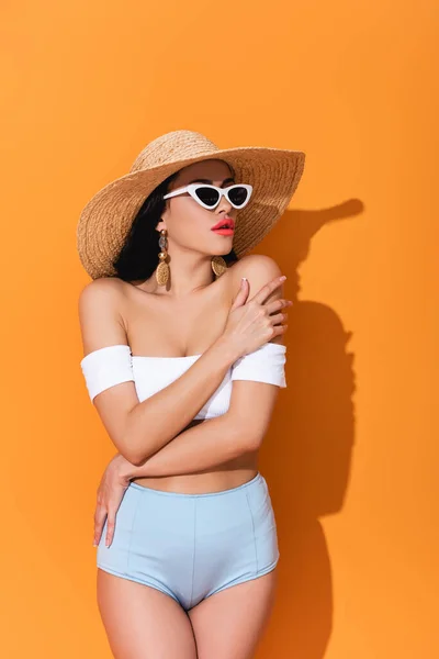 Стильная женщина в купальнике, солнцезащитных очках и соломенной шляпе, стоящая со скрещенными руками на оранжевом — стоковое фото