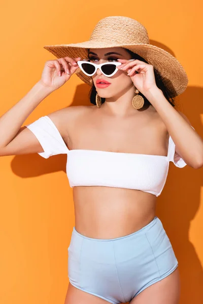 Mujer con estilo en traje de baño y sombrero de paja tocando gafas de sol en naranja - foto de stock