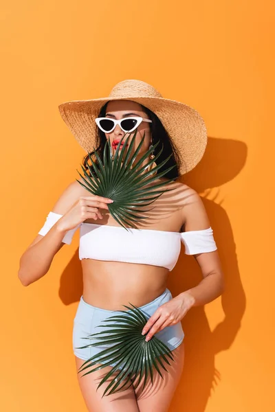 Fashionable woman in swimsuit, sunglasses and straw hat holding palm leaves on orange — Stock Photo