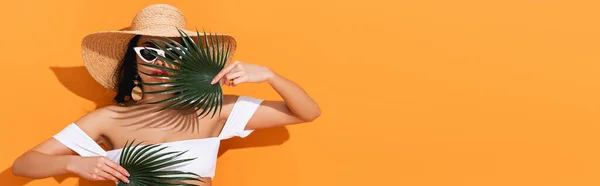 Plan panoramique de femme à la mode en maillot de bain, lunettes de soleil et chapeau de paille tenant des feuilles de palmier sur orange — Photo de stock