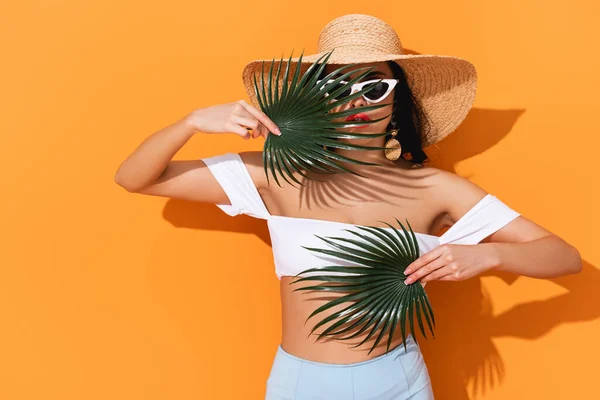 Fashionable girl in swimsuit, sunglasses and straw hat holding palm leaves on orange — Stock Photo