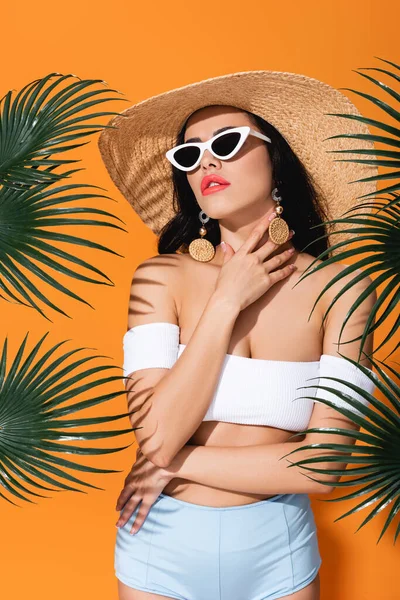 Fashionable girl in swimsuit, sunglasses and straw hat touching neck near palm leaves on orange — Stock Photo