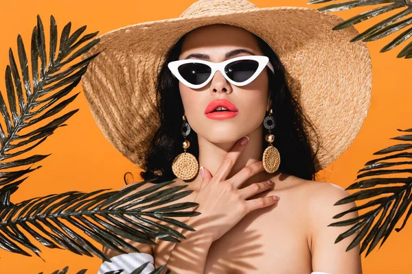 Attraente donna in occhiali da sole e cappello di paglia toccare collo vicino foglie di palma isolato su arancione — Foto stock