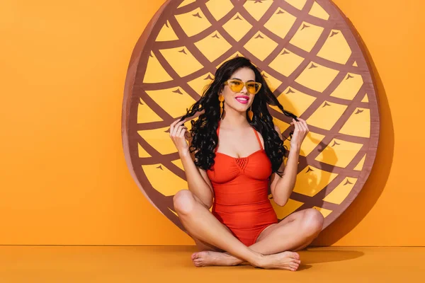 Attractive and barefoot woman in swimsuit and sunglasses sitting in yoga pose near paper cut pineapple on orange, summer concept — Stock Photo