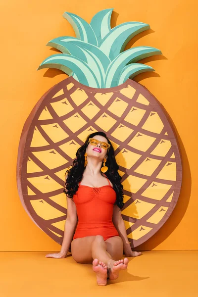 Fröhliche und barfüßige Frau in Badeanzug und Sonnenbrille, die neben papiergeschnittener Ananas auf Orange sitzt, Sommerkonzept — Stockfoto