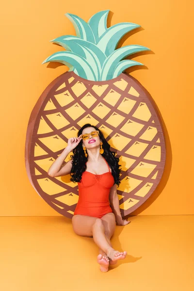 Glückliche und barfüßige Frau im Badeanzug neben papiergeschnittener Ananas und anrührender Sonnenbrille auf orangefarbenem Sommerkonzept — Stockfoto