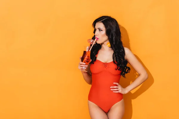 Attractive woman in swimsuit drinking cocktail and standing with hand on hip on orange — Stock Photo