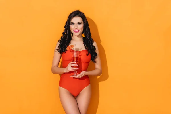 Mujer feliz en traje de baño sosteniendo vaso con cóctel y de pie en naranja - foto de stock