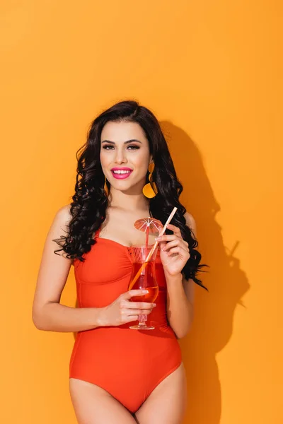 Fröhliche Frau im Badeanzug hält Glas mit Cocktail und steht auf Orange — Stockfoto