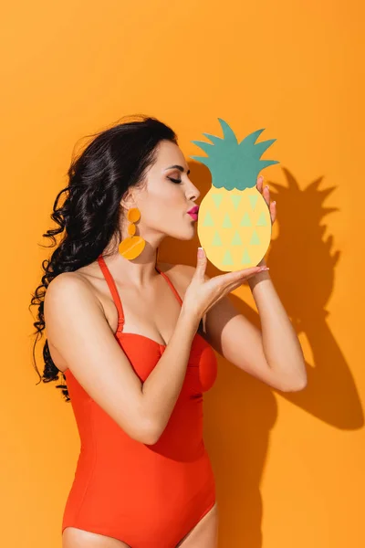 Side view of attractive woman in swimsuit kissing paper cut pineapple on orange — Stock Photo