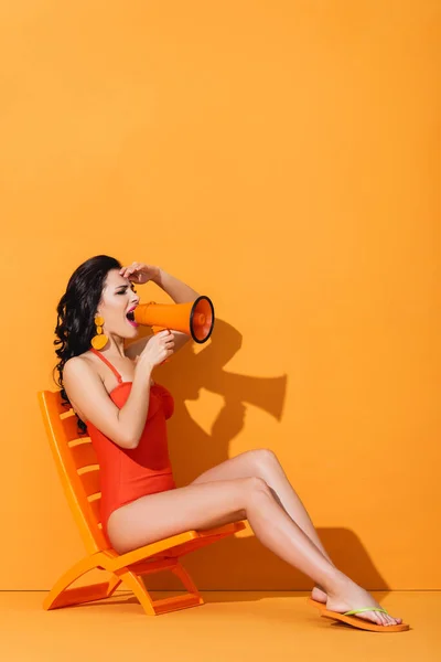 Young woman in swimwear holding megaphone and screaming while sitting on deck chair on orange — Stock Photo