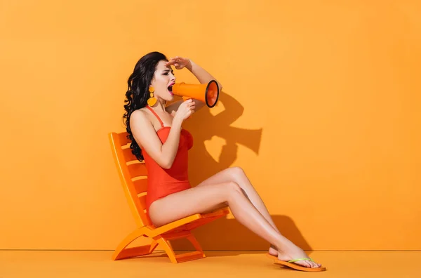 Mulher atraente em roupa de banho segurando megafone e gritando enquanto sentado na cadeira de praia em laranja — Fotografia de Stock