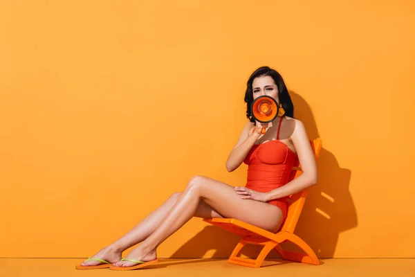 Woman in swimwear holding megaphone and screaming while sitting on deck chair on orange — Stock Photo