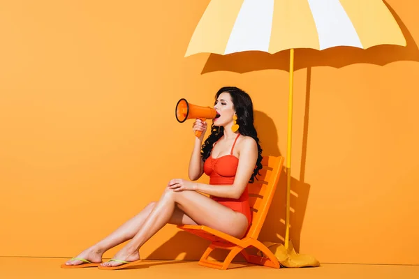 Mujer joven en traje de baño sosteniendo megáfono y gritando mientras está sentado en la silla de cubierta cerca de papel cortado paraguas en naranja - foto de stock