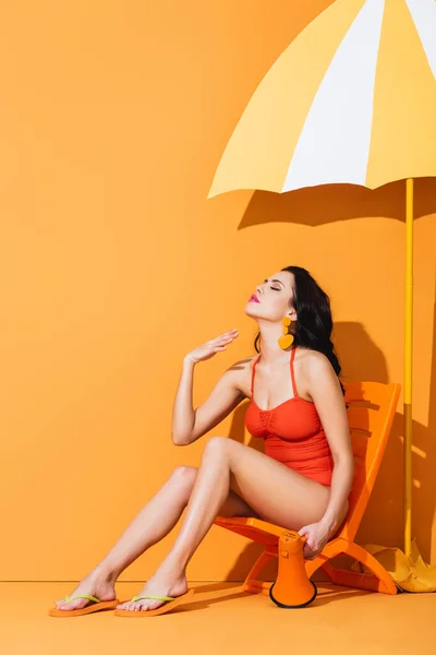 Attractive girl in swimwear holding megaphone while sitting on deck chair and feeling hot near paper cut umbrella on orange — Stock Photo