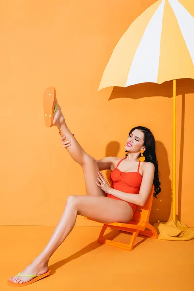 Cheerful young woman in swimsuit touching leg while sitting on deck chair near paper cut umbrella on orange — Stock Photo
