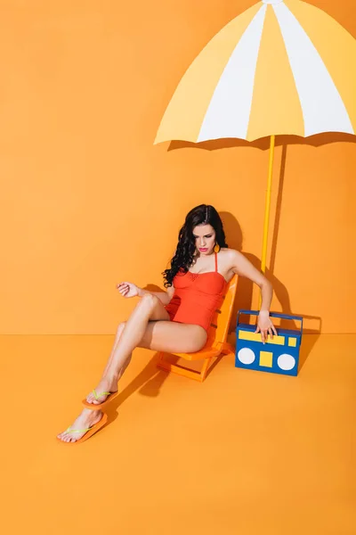 Young woman in swimsuit touching paper boombox while sitting on deck chair near umbrella on orange — Stock Photo